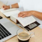 A woman writing in a notebook with coffee and laptop on a wooden desk.